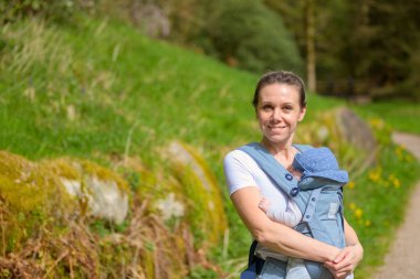 Parktaki bir bebek taşıyıcısında tutarken ve taşırken kameraya bakıp gülümseyen mutlu kadın