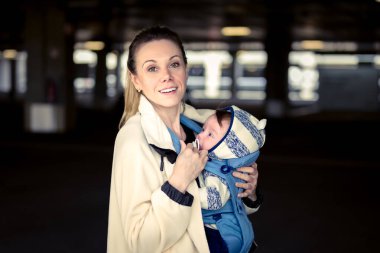 Happy woman looking and smiling to camera while holding and carrying her baby in a baby carrier in a carpark