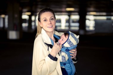 Happy woman looking and smiling to camera while holding and carrying her baby in a baby carrier in a carpark clipart
