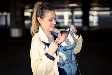 Sideview portrait of a happy woman looking and smiling to her baby while holding and carrying it in a baby carrier in a carpark