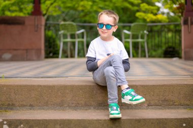 Güneş gözlüğü takan ve günlük kıyafetler giyen genç bir çocuk dışarıda rahat bir şekilde oturuyor..