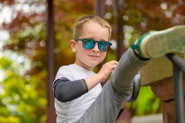 stock image A young boy wearing blue sunglasses, a white and grey shirt, and grey pants is having fun outdoors.