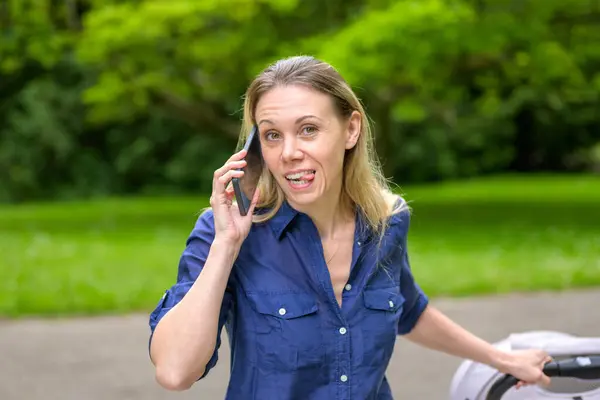 stock image A smiling mother in a blue shirt and white pants enjoys a phone conversation while pushing a stroller through a park.