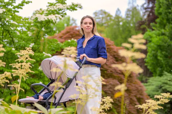 stock image A mother in a blue shirt and white pants walks through a lush park pushing a stroller with her baby.