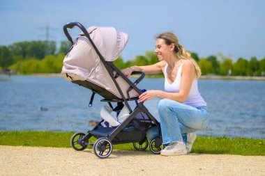 Gülümseyen bir anne göl kenarında güneşli bir günün tadını çıkarırken bebek arabasını ayarlar..