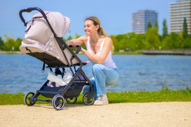 Mutlu bir anne bebek arabasının yanında diz çöker, göl kenarında güzel bir günün tadını çıkarır..