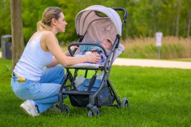 Bir anne, bebeğini bebek arabasıyla gezdirmek için çimenlerin üzerinde diz çöker ve dışarıda huzurlu bir anın tadını çıkarır..