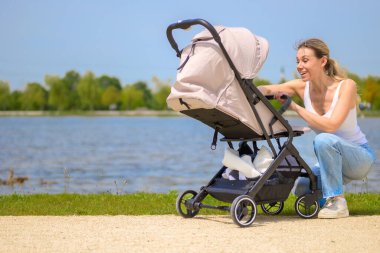 Bir anne, arka planda yüksek binalar olan manzaralı bir parkta bebek arabasının yanında diz çöker..