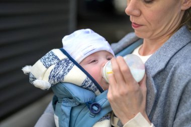 Gri ceketli bir anne tarafından biberondan beslenen bir bebeğin yakın çekimi. Bebek sıcak bir şekilde örgü bir şapka ve ceket giydi.