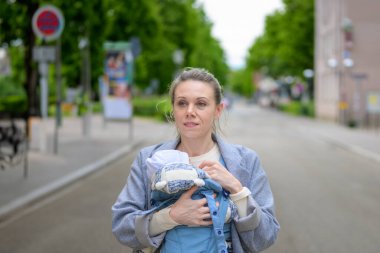 Gri paltolu bir anne güneşli bir caddede yürürken bebeğini mavi bir bebek taşıyıcısının içinde taşıyor. Bebek sıcak bir şekilde örgü bir şapka ve ceket giydi..