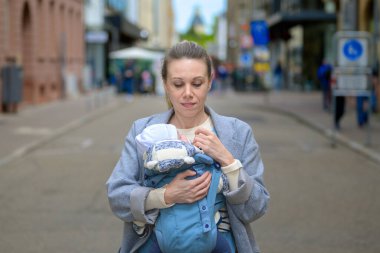 Gri paltolu bir anne, bebeğini mavi bir bebek taşıyıcısının içinde şehir caddesinde dururken taşıyor. Bebek sıcak bir şekilde örgü bir şapka ve ceket giydi.
