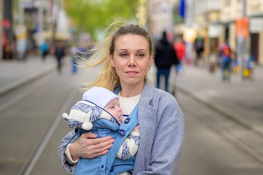 Gri paltolu bir anne, bebeğini işlek bir caddede dururken mavi bir bebek taşıyıcısının içinde taşıyor. Bebek sıcak bir şekilde örgü bir şapka ve ceket giydi.