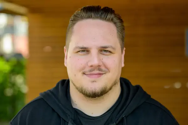 stock image A young man with neatly combed hair and a trimmed beard smiles confidently at the camera. He is wearing a black hoodie and standing in front of a wooden background, giving a casual and relaxed vibe.