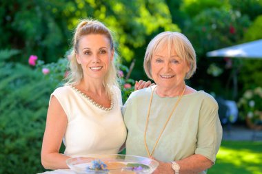 Gülümseyen yaşlı bir kadın ve genç bir kadın birlikte poz veriyorlar, yemyeşil ve çiçeklerle dolu yemyeşil bir bahçede güneşli bir günün tadını çıkarıyorlar..