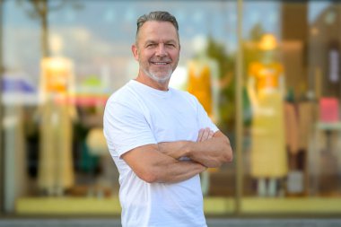 Orta yaşlı bir adam kollarını kavuşturmuş, rahat beyaz bir tişört giyiyor..