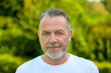 Beyaz tişört giyen, gri saçlı ve sakallı olgun bir adamın yakın plan portresi..