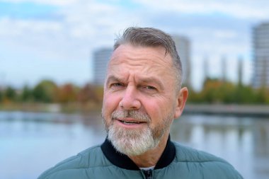 Gri sakallı olgun bir adamın yakın plan portresi. Meraklı ve az da olsa eğlendirici bir ifade sergiliyor..