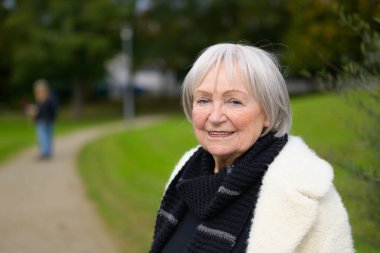 Yaşlı bir kadın, kısa, gri saçlı ve sıcak, nazik bir gülümsemesi olan bir park yolunda duruyor..