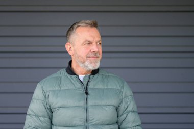 Gri sakallı orta yaşlı bir adam açık yeşil bir kabarık ceket giymiş düşünceli bir ifadeyle yan tarafa bakıyor..