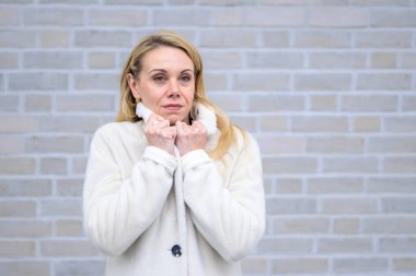 Yumuşak beyaz önlüklü sarışın bir kadın yakasını sıkı sıkı tutuyor. Yüzü sakin ve içten içe bakıyor..