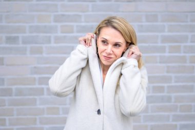 Yumuşak beyaz bir ceket giymiş sarışın bir kadın nazikçe yakasını tutuyor, düşünceli ve düşünceli bir tavır sergiliyor..