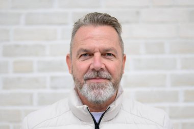 Gri sakallı, nazik gülümsemeli, doğrudan kameraya bakan olgun bir adamın portresi..