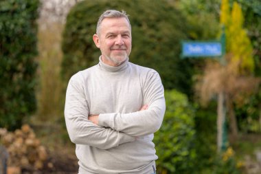 Orta yaşlı, kısa gri saçlı ve bakımlı sakallı bir adam açık hava bahçesinde çapraz kollarıyla duruyor..