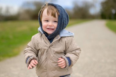 Bej renkli örtülü bir ceket giyen neşeli bir genç çocuk ve çakıl yolunda dururken tüylü mavi bir başlık gülümsüyor..