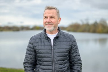 Koyu renk yastıklı ceketli bir adam huzur dolu bir gölün yanında duruyor, yukarı bakarken hafifçe gülümsüyor..