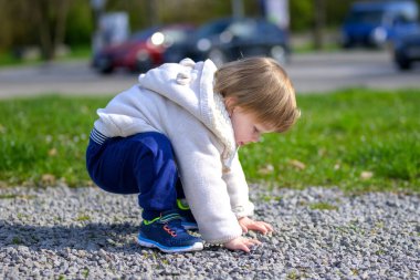 Küçük bir çocuk çakıllı bir yolda çömelmiş, iki eliyle küçük taşlar toplamaya odaklanmış..