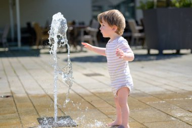 Genç bir çocuk, yer seviyesindeki bir çeşmenin önünde çıplak ayakla durup merakla uzanır..