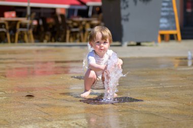 Çizgili sandalyede oturan genç bir çocuk halka açık bir meydandaki çeşmenin üzerine tuhaf bir şekilde çömeliyor..