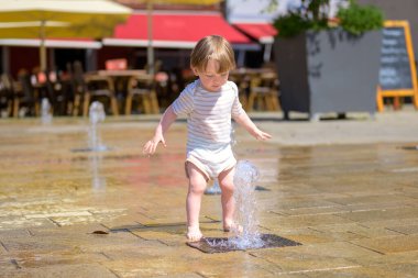 Çizgili kemerli küçük bir çocuk kollarını açmış güneşli bir meydanda yükselen bir su jetinin üzerinde duruyor..