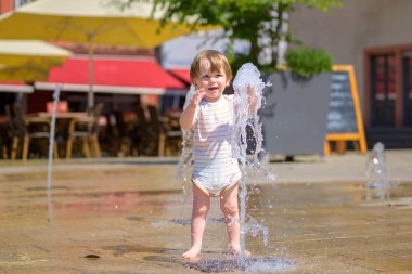 Çizgili bir sandalyede mutlu bir çocuk güneşli bir günde halka açık bir fıskiyede su sıçratıyor..