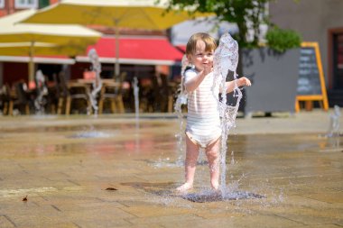 Çizgili kemerli küçük bir çocuk güneşli bir yaz gününde halka açık bir su çeşmesiyle yalınayak oynuyor..
