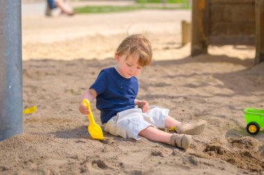 Küçük bir çocuk kum havuzunda tek başına oturuyor, elinde sarı bir oyuncak kürekle yere odaklanmış..