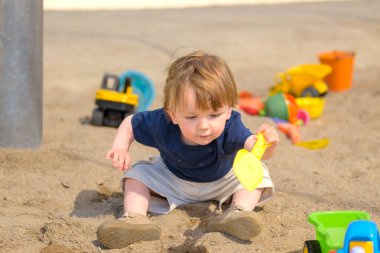 Küçük bir çocuk, elinde sarı bir plastik kürekle bir kum havuzunda dikkatlice oynar ve kazarken öne doğru eğilir..