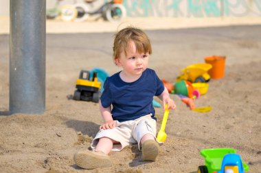 Güneşli bir kum havuzunda elinde sarı bir kürekle oturan bir çocuk düşünceli bir şekilde uzaklığa bakıyor..