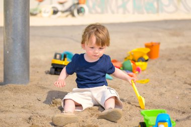 Genç bir çocuk güneşli bir günde kum havuzunda oturuyor, elinde sarı plastik bir kürek ve kumu kazıyor..