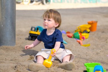 Güneşli bir günde kum havuzunda oturan bir çocuk elinde sarı bir oyuncak kürekle renkli kum oyuncaklarıyla çevrili..