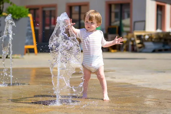 Çizgili tulumlu küçük çocuk şehir meydanında bir çeşmeyle oynuyor..