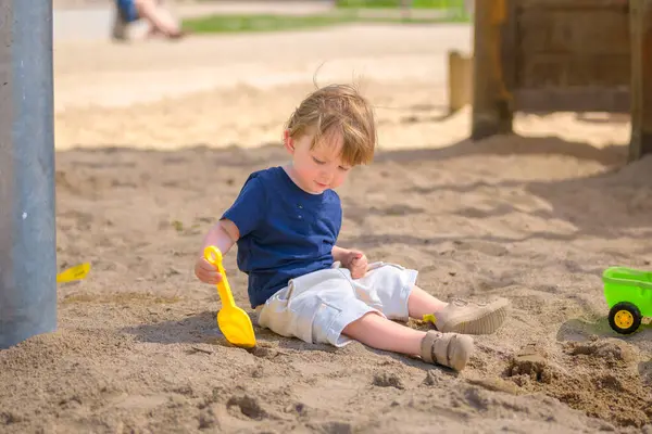 Küçük bir çocuk kum havuzunda tek başına oturuyor, elinde sarı bir oyuncak kürekle yere odaklanmış..