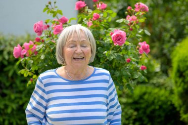 Mavi ve beyaz çizgili gömlekli neşeli yaşlı kadın. Gözleri kapalı gülüyor. Çiçek açan pembe güllerin ve yemyeşil çiçeklerin önünde duruyor..