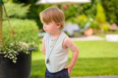 Kolsuz çizgili gömlekli ve koyu şortlu genç bir çocuk yemyeşil bir bahçede sessizce duruyor..