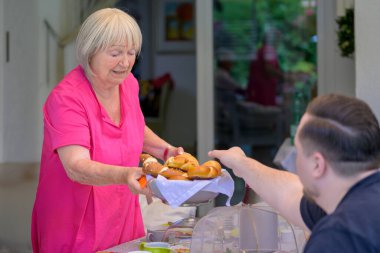 Açık pembe bluzlu yaşlı bir kadın, açık hava aile toplantısı sırasında oturan bir adama geleneksel kraker sepeti sunuyor..