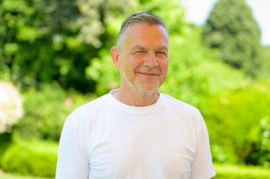 Kendinden emin yaşlı bir adam beyaz bir tişört giyiyor ve güneşli bir günde dışarıda dururken nazikçe gülümsüyor..