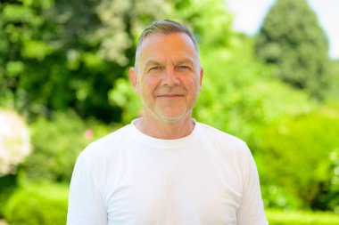 Beyaz tişörtlü yaşlı bir adam sakince dışarıda, açık yeşil bir park ortamında dikiliyor..