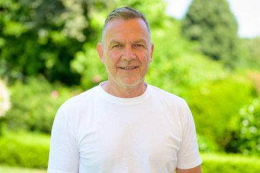 Beyaz tişörtlü yaşlı bir adam güneşli bir bahçede gülümsüyor..