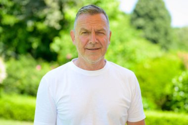 Sade beyaz tişört giyen kendinden emin yaşlı bir adam açık havada, yemyeşil bir bahçede parlak bir yaz gününde duruyor..