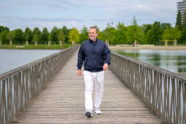 Normal bir park ortamında tahta bir köprüde güvenle yürüyen yaşlı bir adam..